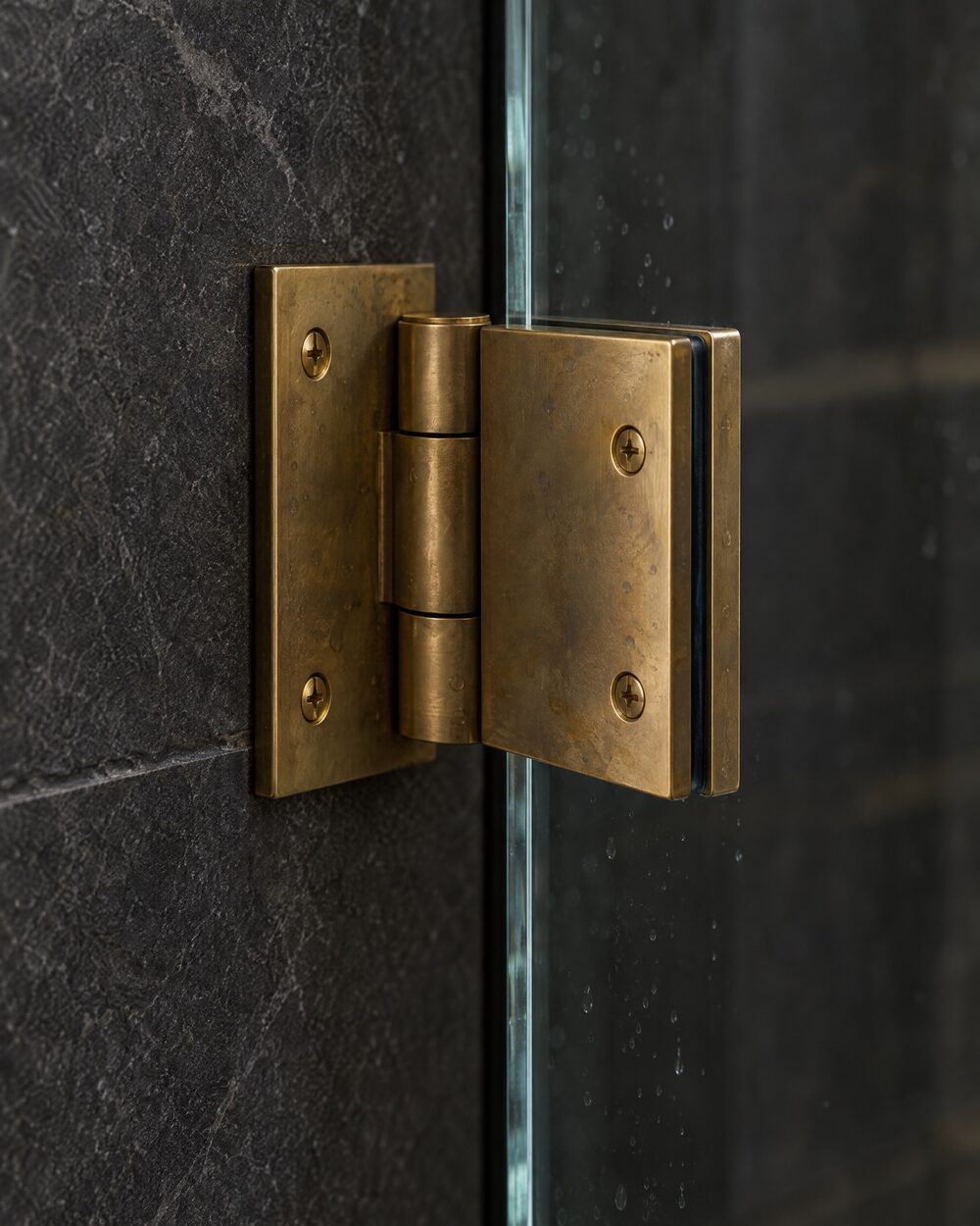 Close-up of a brass glass-to-wall hinge holding the edge of a clear frameless shower door against dark stone tile.