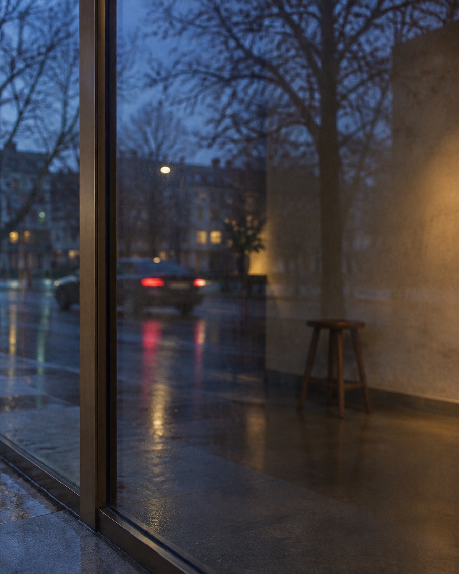 A shop window at dusk reflecting a wet street, with a dim plaster interior behind.