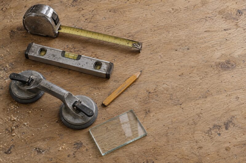 A tape measure, spirit level, glass suction lifter, pencil and a small offcut of clear glass on an oak bench.